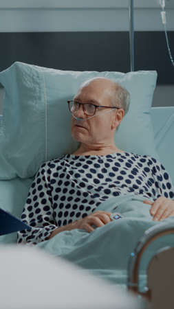 Multi ethnic medical staff explaining disease to patient in hospital ward bed at clinic. Sick old man with nasal oxygen tube in intensive care room preparing for medicine against health problemsの写真素材