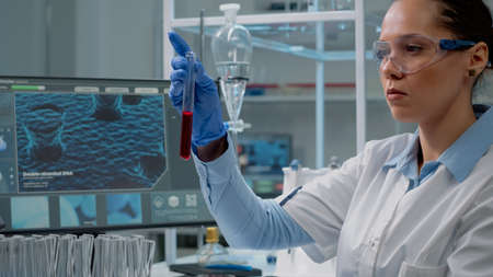Medicine scientist using computer while holding test tube with red liquid solution for research...