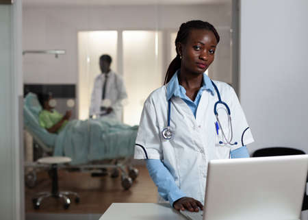 African american doctor looking at camera in hospital ward with medical equipment and instruments. Portrait of african ethnicity woman with stethoscope and white coat at clinic deskの写真素材