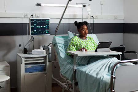 African american patient sitting in hospital ward bed with digital tablet chatting and web surfing. Young person with illness waiting on diagnosis advice while playing with online technologyの写真素材