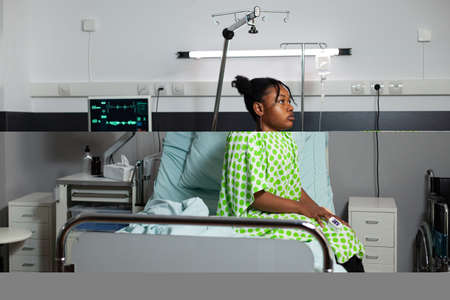 Sick patient of african american ethnicity laying on bed in hospital ward. Black teenager sitting...