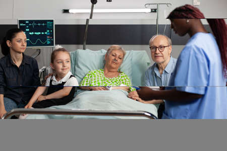 African american asisstant analyzing medical expertise explaining sickness treatment to elderly sick woman. Caring family checkup retired patient to support her during clincal therapy in hospital wardの写真素材