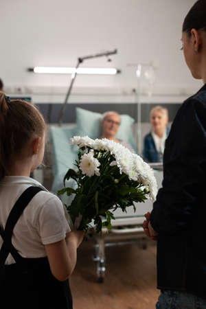 Caring granddaughter bringing flowers gift to sick old grandfather during clinical recovery in hospital ward. Elderly senior man resting in bed recovering after medical surgery. Healthcare supportの写真素材