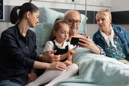 Cheerful family visiting elderly grandfather in hospital ward during clinical recovery waiting for...