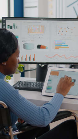 Black handicapped woman entrepreneur with locomotor disabilities using computer and tablet in same time working in business start-up office sitting in wheelchair. Busy multitasking african employeeの写真素材