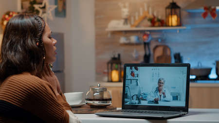 Woman with flu using telemedicine on laptop at home in christmas decorated kitchen. Sick young adult...