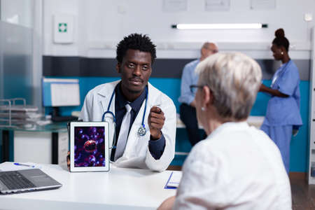 Doctor of african american ethnicity holding digital tablet with virus animation while sitting at desk. Black medic explaining disease bacteria and healthcare problems to old patientの写真素材
