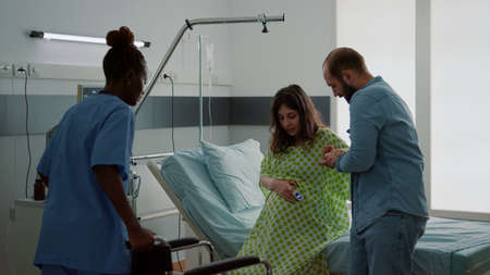 African american nurse helping pregnant wife sit in wheelchair at maternity ward in hospital. Young...
