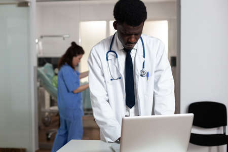 Portrait of african ethnicity doctor standing at desk with laptop in hospital ward at clinic....