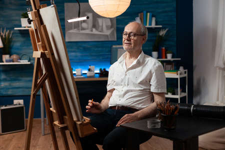 Caucasian senior man working as artist with canvas and easel for drawing masterpiece with pencil. Older artist sitting in artwork studio creating fine art as creative hobby indoorsの写真素材