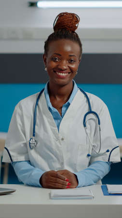 Portrait of woman working as medic in office at healthcare clinic. Doctor wearing white coat and stethoscope while sitting at desk in medical cabinet smiling and looking at cameraの写真素材