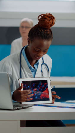 Black doctor holding virus animation on tablet showing disease cells to elder patient at desk. African american medic explaining coronavirus bacteria using modern technology on device.の写真素材