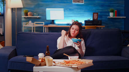POV of adult sitting on couch laughing while looking at TV and eating chips from bowl. Caucasian woman having fun watching television after work with takeaway food on table in living roomの写真素材
