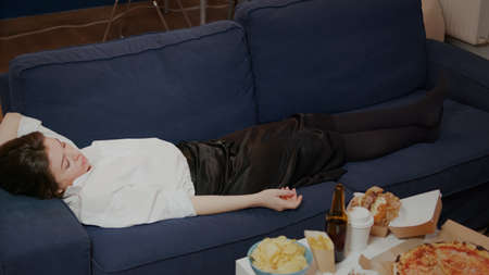 Young person falling asleep on couch dropping TV remote at home. Tired woman from work sleeping after eating takeaway food in living room. Adult taking nap while watching televisionの写真素材