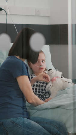 Hospitalized sick child resting in bed wearing nasal tube recovering after medical surgery during sickness examination in hospital ward. Mother discussing medication treatment for health recoveryの写真素材