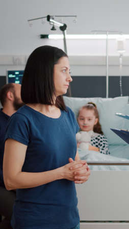 Mother discussing medical treatment with pediatrician doctor during disease examination in hospital ward. Girk kid resting in bed while african american nurse writing sickness diagnosis on clipboardの写真素材