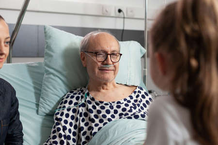 Selectiv focus of elderly sick old man resting in bed talking with caring granddaughter after medical expertise hospital ward. Grandfather enjoying spending time with child during clinical surgeryの写真素材