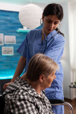 Support nurse worker listening lungs of retired female in wheelchair using medical stethoscope during examination. Social services nursing elderly retired female. Healthcare assistanceの写真素材