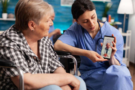 Social worker holding smartphone for disabled senior retired woman in wheelchair during online medical video call teleconference with physician doctor. Healthcare treatment in nursing homeの写真素材