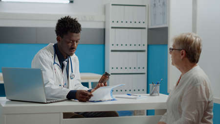 Young man working as medic holding bottle of pills for prescription treatment against disease. Doctor giving sick elderly patient flask of medicine, drugs and medicament in cabinetの写真素材