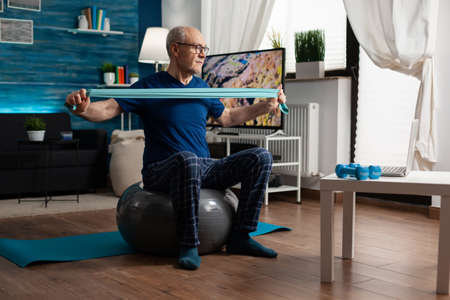 Invalid pensioner practicing active arm exercises using resistance elastic band watching online fitness workout video on laptop. Pensioner working body healthcare sitting on swiss ball in living roomの写真素材