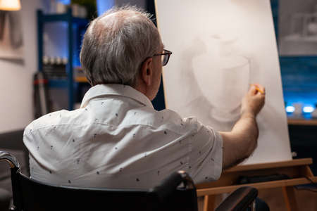 Senior artist with handicap creating vase drawing on canvas with pencil in artwork studio. Caucasian old disabled man sitting in wheelchair while working on professional masterpieceの写真素材