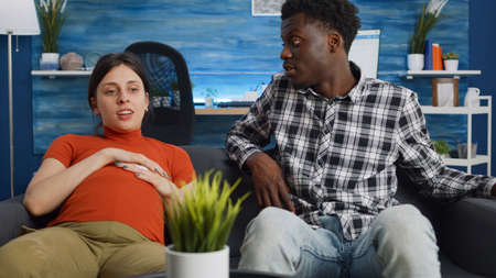 Interracial couple talking about child and parenthood while sitting on couch at home. Mixed race...