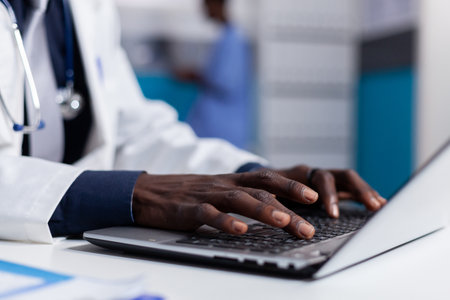 Close up of black hands typing on laptop keyboard while sitting at white desk in medical office. African american man wearing medical coat using computer device and modern technologyの写真素材