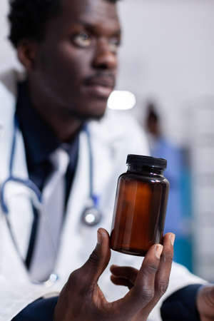 Close up of african american hand with bottle of pills for treatment recommendation. Black doctor holding flask with medicine, medicament and drugs. Medic with vial in medical cabinetの写真素材