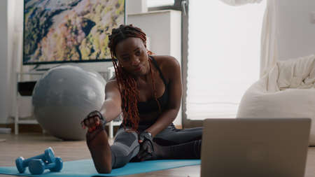 Woman with black skin doing workout in living room stretching exercise on yoga map while watching fitness video on laptop. Flexible adult training body muscle enjoying healthy lifestyleの写真素材