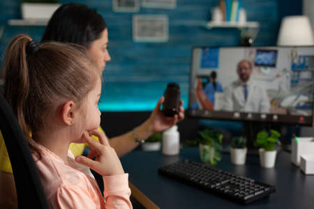 Concerned mother talking to doctor on video call conference technology about child healthcare problems. Caucasian adult using telemedicine for healing treatment diagnosis and pillsの写真素材