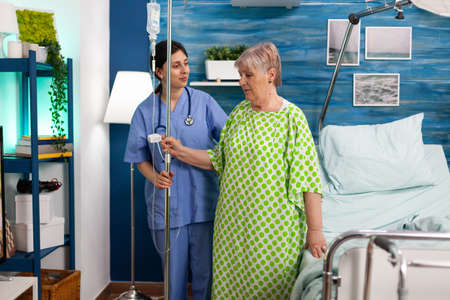 Support assistant worker caring disabled retired senior old woman with IV drip bag during medical recovery. Social services nursing elderly retired female. Healthcare assistance. Caregiver nurseの写真素材