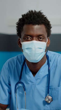 Portrait of man with nurse occupation looking at camera with face mask and uniform. Medical assistant sitting at desk in healthcare office with laptop and professional instrumentsの写真素材