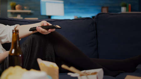 Close up of caucasian hand holding television remote while person laying on couch in living room. Woman relaxing on sofa at home with takeaway meal of fast food for unhealthy dinnerの写真素材