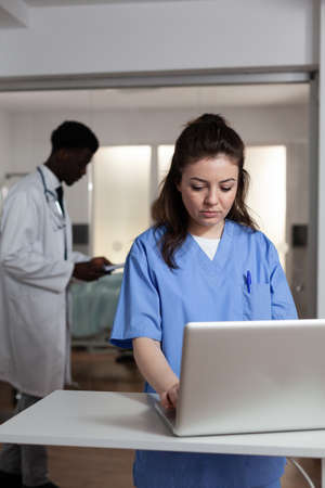 Caucasian woman using laptop at desk in hospital ward for professional healthcare. Young adult with nurse occupation working as consultant for specialist, surgeon, medic, doctorの写真素材