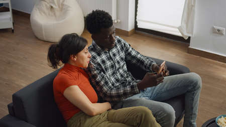 Married interracial couple taking selfie with smartphone while sitting together on sofa at home....