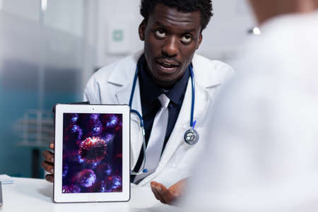 Doctor of african american ethnicity holding digital tablet with virus animation while sitting at...