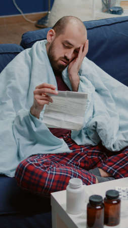 Man with sickness reading medicament information leaflet to heal cold and flu. Person with virus symptoms holding medication paper against disease in blanket while having headache.の写真素材