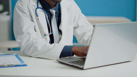 Close up of doctor typing on laptop keyboard in medical cabinet at healthcare facility. Portrait of medic sitting at desk with documents using device with technology for consultationsの写真素材