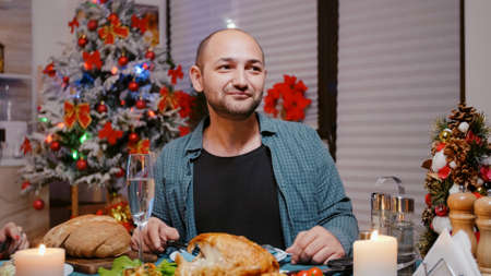 Close up of man talking on video call and waving while eating chicken with woman at festive dinner. Adult using online conference for remote communication, celebrating christmas eve.の写真素材