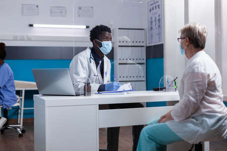 African american doctor sitting at desk talking to old woman about medical treatment for disease. Black medic consulting elderly patient wearing face masks during coronavirus pandemicの写真素材