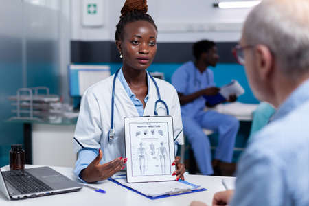 African american doctor holding illustration on tablet screen showing results to elder patient at...