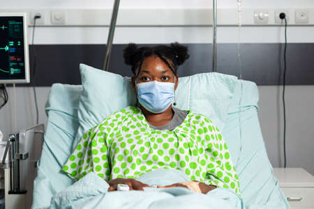 Portrait of african american patient with face mask looking at camera sitting in hospital ward bed. Young woman with disease, illness waiting on consultation with medical equipmentの写真素材