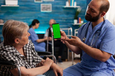 Man social worker looking at mock up green screen chroma key with isolated display while nursing disabled senior woman at home. Social services nursing elderly retired female. Support assistantの写真素材