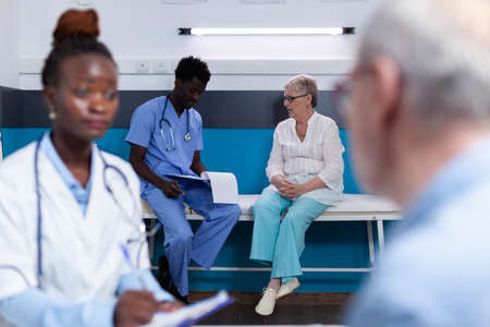 Black medic and caucasian old patient talking sitting at desk in medical cabinet. African american man with nurse profession consulting elder woman on bed in background at clinicの写真素材