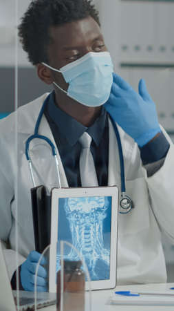 Healthcare specialist holdind x ray on tablet explaining scan examination results to old woman at desk with plexiglass wall. Doctor with face mask looking at radiography on digital deviceの写真素材