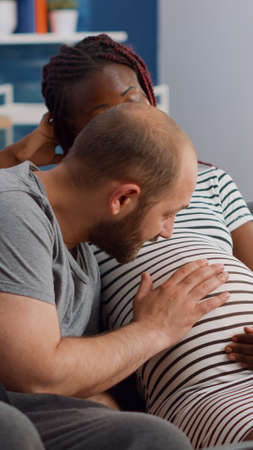 Interracial couple expecting child in living room. Caucasian husband talking to baby and touching belly while african american wife relaxing on couch at home. Mixed race partnersの写真素材