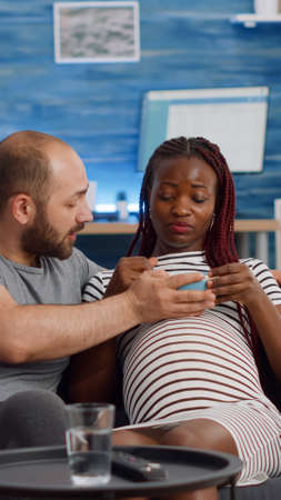 Interracial couple with pregnancy watching movie on TV while eating popcorn. Multi ethnic parents expecting baby and sitting on living room couch. Mixed race people relaxing with televisionの写真素材