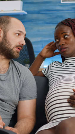 Interracial couple expecting child and bonding on couch at home. Mixed race people touching belly with baby and talking about parenthood. Multi ethnic parents with pregnancy.の写真素材