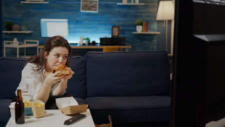 Caucasian woman eating hamburger from delivery bag while watching movie on television at home. Adult enjoying takeaway fast food meal and bottle of beer sitting on couch in living roomの写真素材